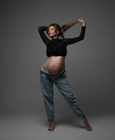 a pregnant woman poses in jeans and a black top with her hands behind her head