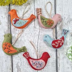 four felt bird ornaments hanging from clothes pins on a white wooden surface with flowers in the background