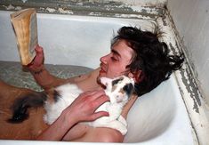 a man laying in a bathtub holding a cat and reading a book with his hand