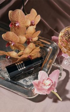 an orchid and lipstick on a tray next to a glass vase with flowers in it