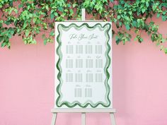 a wedding seating chart on an easel in front of a pink wall with green leaves