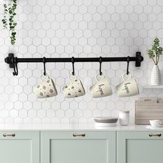 four coffee mugs hanging on a black bar in a kitchen with white hexagonal tiles