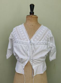 Early 20th Century original lace trimmed camisole top / corset cover. Deep V necked and with short sleeves.  Fastens down the front with two cloth covered laundry buttons and has a  small peplum underneath.   The waist ties with a drawstring that can be adjusted. The deep V neckline is flanked by strips of broderie anglaise interspersed with netted lace to form a chevron motif yoke.  The neck is adjustable with the fine ribbon.  Ribbon insert lace runs across the front of the camisole marking th Cottagecore Short Sleeve Top With Lace Collar, Fitted Cropped Top With Lace Collar, Fitted Broderie Anglaise Short Sleeve Top, Fitted Short Sleeve Top With Broderie Anglaise, Fitted Victorian Tops With Lace Collar, Victorian Lace Trim Top For Daywear, Summer Cotton Tops With Lace Work, Cotton Lace Work Tops For Summer, Vintage Fitted Tops With Broderie Anglaise