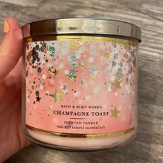 a hand holding a pink candle with gold and white stars on the lid that says bath & body works champagne toast