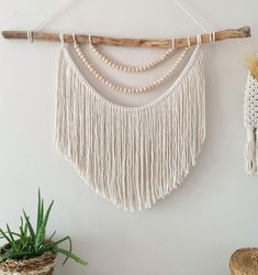 a white wall hanging with beads and tassels next to a potted plant