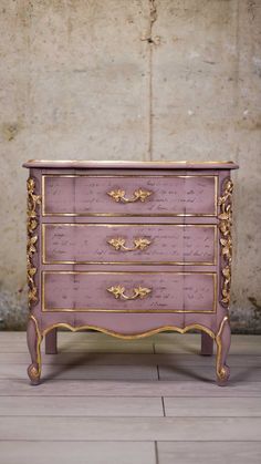 a pink and gold chest of drawers with writing on the drawer top, in front of a stone wall