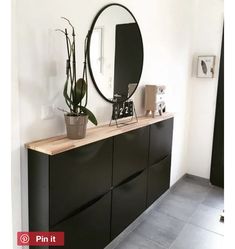 a black dresser with mirror and plants on it in a white walled room, next to a planter