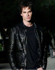 a young man in a black shirt and leather jacket standing on the street at night