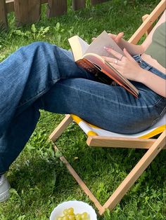 a person sitting in a lawn chair with a book and bowl of fruit on the grass