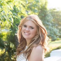 a beautiful young woman standing in front of a white car smiling at the camera with trees and bushes behind her