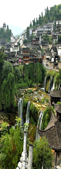 a river running through a lush green hillside covered in forest next to tall buildings and trees