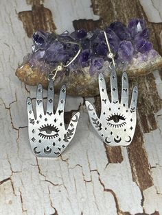 Palmistry hand earrings are a unique and eye-catching piece of jewelry thats. featuring two small hands made of metal. The hands are designed to resemble the hands of a palmist. they are adorned with small symbols or charms, such as moons or stars and the evil eye for protection, which add to their mystical appeal. These earrings offer a spiritual look, making them perfect for those who are interested in palmistry, astrology, or other forms of divination.  They are sure to start conversations and spark intrigue. These earrings are a unique and meaningful addition to any jewelry collection and make a great gift for those interested in alternative spirituality and symbolism. Symbolic Silver Dangle Jewelry, Silver Symbolic Hypoallergenic Earrings, Silver Hypoallergenic Symbolic Earrings, Silver Symbolic Earrings For Jewelry Making, Symbolic Silver Earrings With Ear Wire, Symbolic Silver Pierced Earrings, Symbolic Silver Jewelry With Matching Earrings, Adjustable Hand Cast Earrings As Gift, Symbolic Handmade Dangle Jewelry