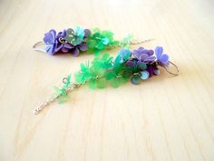 two purple and green flowers are hanging from silver earwires on a wooden surface