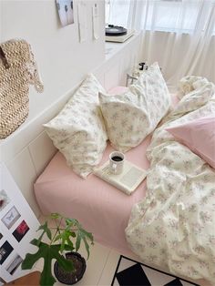 a bed with pink sheets, pillows and a book on it next to a potted plant