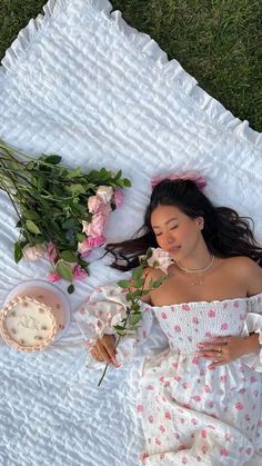 a woman laying on top of a white blanket next to flowers and a bowl of cake