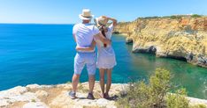 a man and woman standing on top of a cliff next to the ocean looking at the water