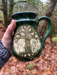 a hand holding a green ceramic mug with an owl design on the side and trees in the background