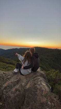 two people sitting on top of a large rock pointing at the sun setting in the distance