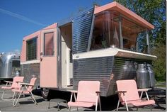 a pink and silver trailer parked in a parking lot next to two lawn chairs on the ground