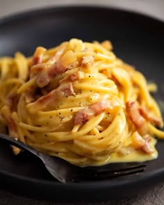 a black plate topped with pasta covered in cheese and bacon next to a silver fork