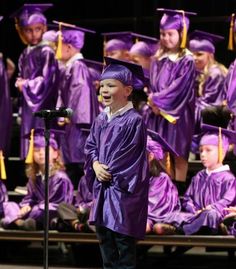 a young boy in purple graduation gown standing on stage with other graduates and microphones behind him