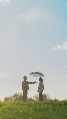 two people standing under an umbrella on top of a green hill with trees in the background