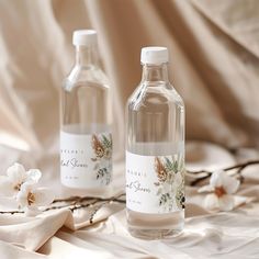 two water bottles with labels on them sitting on a bed next to some white flowers