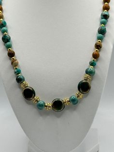 The beads are predominantly in shades of turquoise and brown with a glossy finish, and some have a marbled effect that adds depth and interest to their appearance. Between the larger beads are ornate gold-toned spacer beads with intricate designs, contributing to the necklace's luxurious look. The combination of the earthy brown tones with the vibrant turquoise creates a visually striking contrast. This necklaceswould appeal to those with a taste for bold, statement jewelry that combines natural Artisan Turquoise Necklace With Polished Gold Beads, Artisan Gold Turquoise Necklace With Polished Beads, Brown Beaded Turquoise Necklace With Round Beads, Elegant Turquoise Agate Beaded Necklaces, Turquoise Agate Round Beads Gems And Cabochons, Turquoise Agate Round Beads, Gems And Cabochons, Turquoise Agate Round Beads For Crafting, Turquoise Agate Necklace With Colorful Beads, Chrysocolla Beaded Necklaces For Jewelry Making