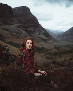 a woman sitting on top of a grass covered hillside next to a valley with mountains in the background