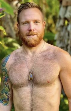 a shirtless man with tattoos on his chest standing in front of trees and bushes