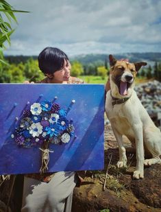 a dog is sitting next to a woman holding a painting that has flowers on it