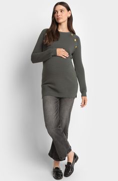 a pregnant woman standing in front of a white wall wearing grey jeans and a green sweater