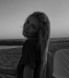 a woman sitting on top of a sandy beach next to the ocean with her hair blowing in the wind