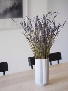 a white vase filled with purple flowers sitting on top of a wooden dining room table