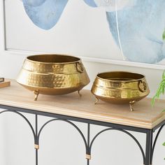 two golden bowls sitting on top of a wooden table next to a green plant in front of a blue and white painting