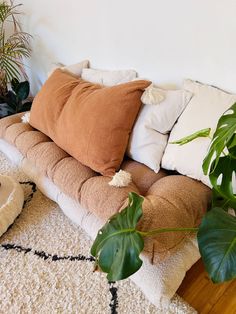 a couch with many pillows on it in front of a wall and some potted plants