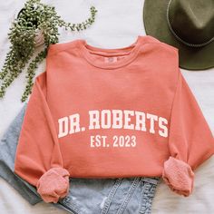 an orange sweatshirt with the words dr roberts est 2023 on it next to a hat and jeans