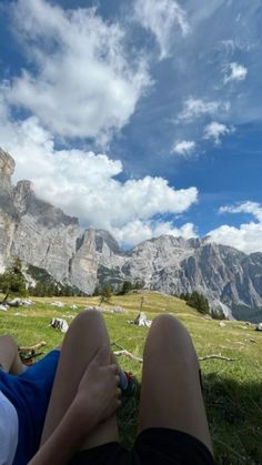 two people laying on the grass in front of some mountains with clouds and blue sky