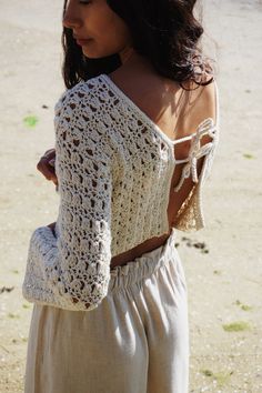 a woman standing on top of a sandy beach wearing a white crochet sweater