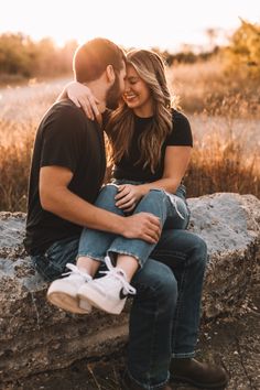 a man and woman are sitting on a rock in front of some dry grass while the sun is setting