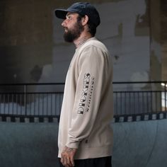 a man with a beard wearing a tan sweatshirt and black pants standing in front of a skateboard ramp