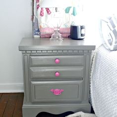 a gray dresser with pink knobs in a white room next to a bed and window