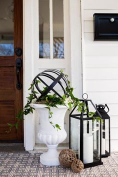 a white vase filled with greenery next to two lanterns on the front step of a house