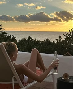 a woman sitting in a lawn chair next to the ocean with a bottle of wine