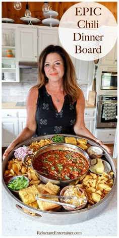 a woman holding a platter full of food with the words epic chili dinner board