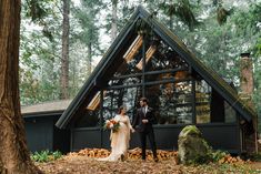 a bride and groom standing in front of a cabin at their wedding ceremony, surrounded by trees