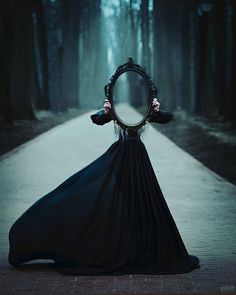 a woman in a black dress holding a mirror over her head while standing on a path