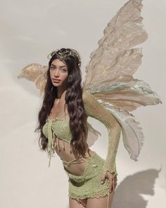 a beautiful woman in a green costume with wings on her head and body, standing next to a white wall