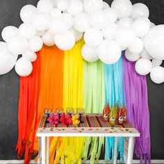 a table topped with lots of balloons next to a rainbow colored wall filled with white balloons