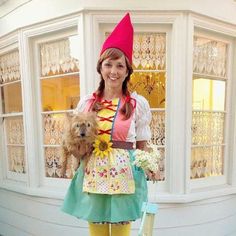 a woman in a costume holding a dog and posing for the camera with a house behind her